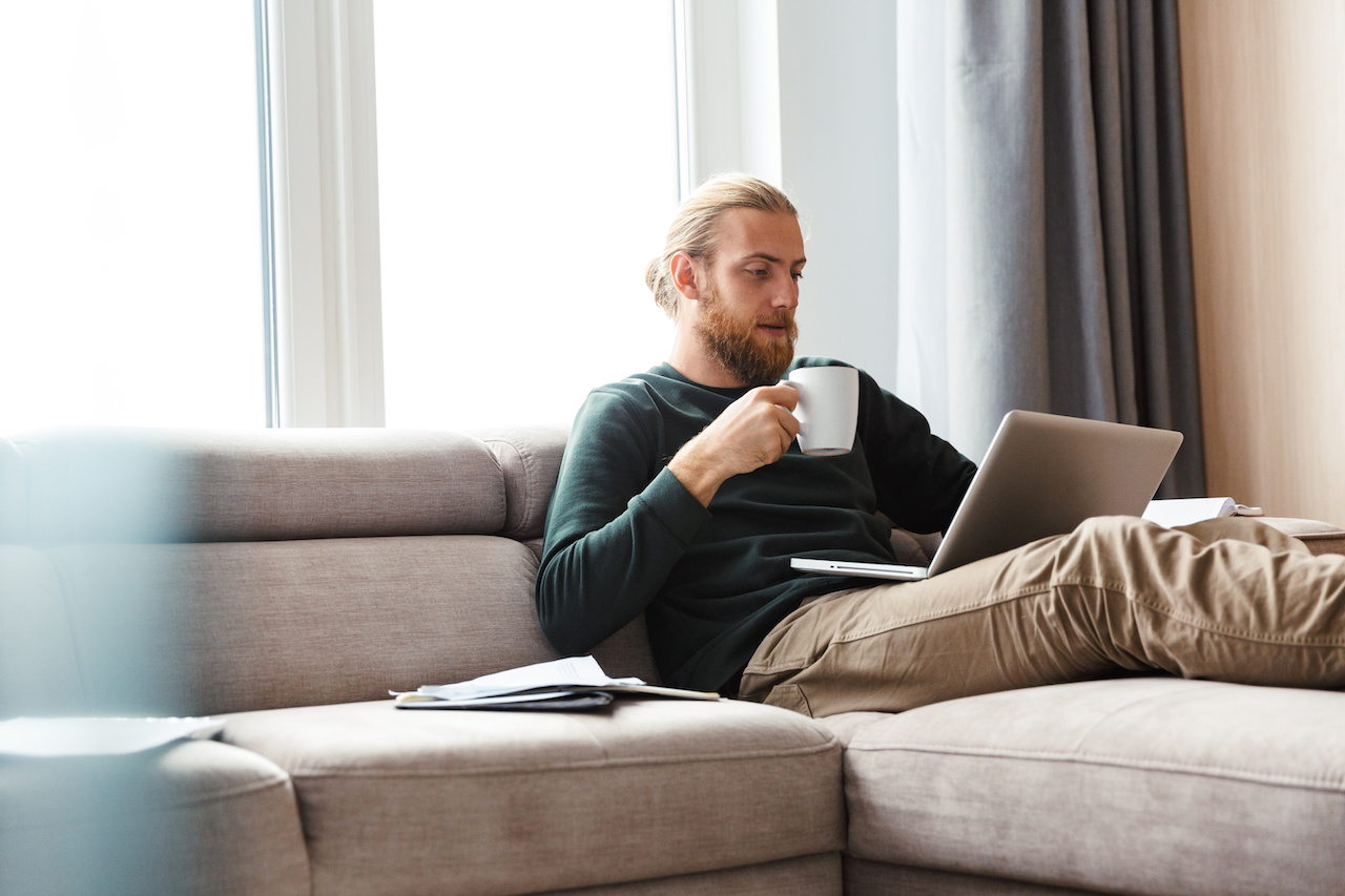 Image of a concentrated young bearded man sitting in home using laptop computer.