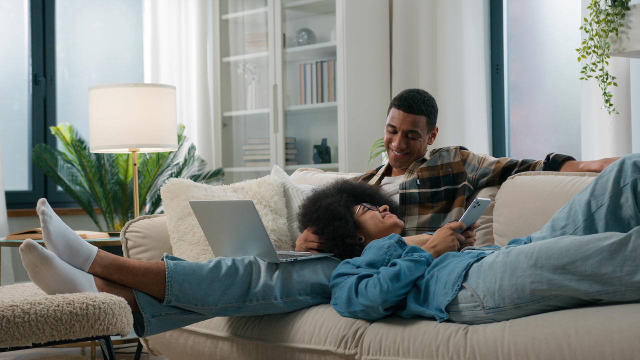 Chilling African American relaxed couple on couch at home together gadget internet addict man working using laptop woman laying browsing mobile phone at apartment spouses social media addiction online