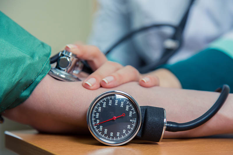 A healthcare professional measures a patient's blood pressure using a manual sphygmomanometer. The stethoscope is placed on the patient’s inner arm while the gauge, showing the pressure reading, rests on the table in clear view.