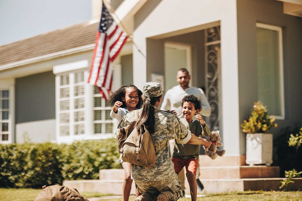 A female soldier in uniform kneels to embrace her smiling children outside their home, as a man watches happily from the doorway. An American flag is displayed near the entrance, symbolizing patriotism and family reunion.