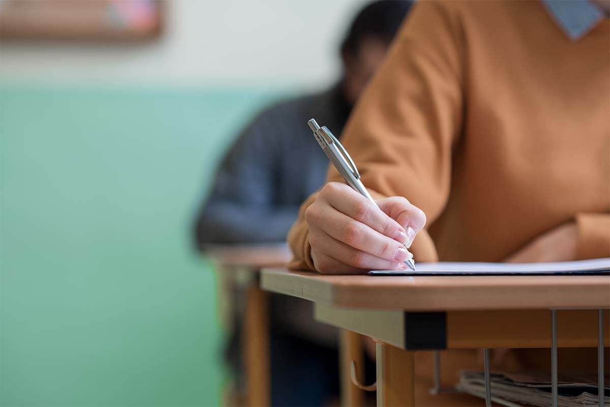 AdobeStock_197909091 Student writes with a pen at a desk in a classroom, with other students blurred in the background.