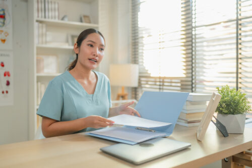 Female healthcare professional reviewing medical documents at desk with laptop in clinic office, administrative medical workflow, patient record management and modern healthcare documentation concept.